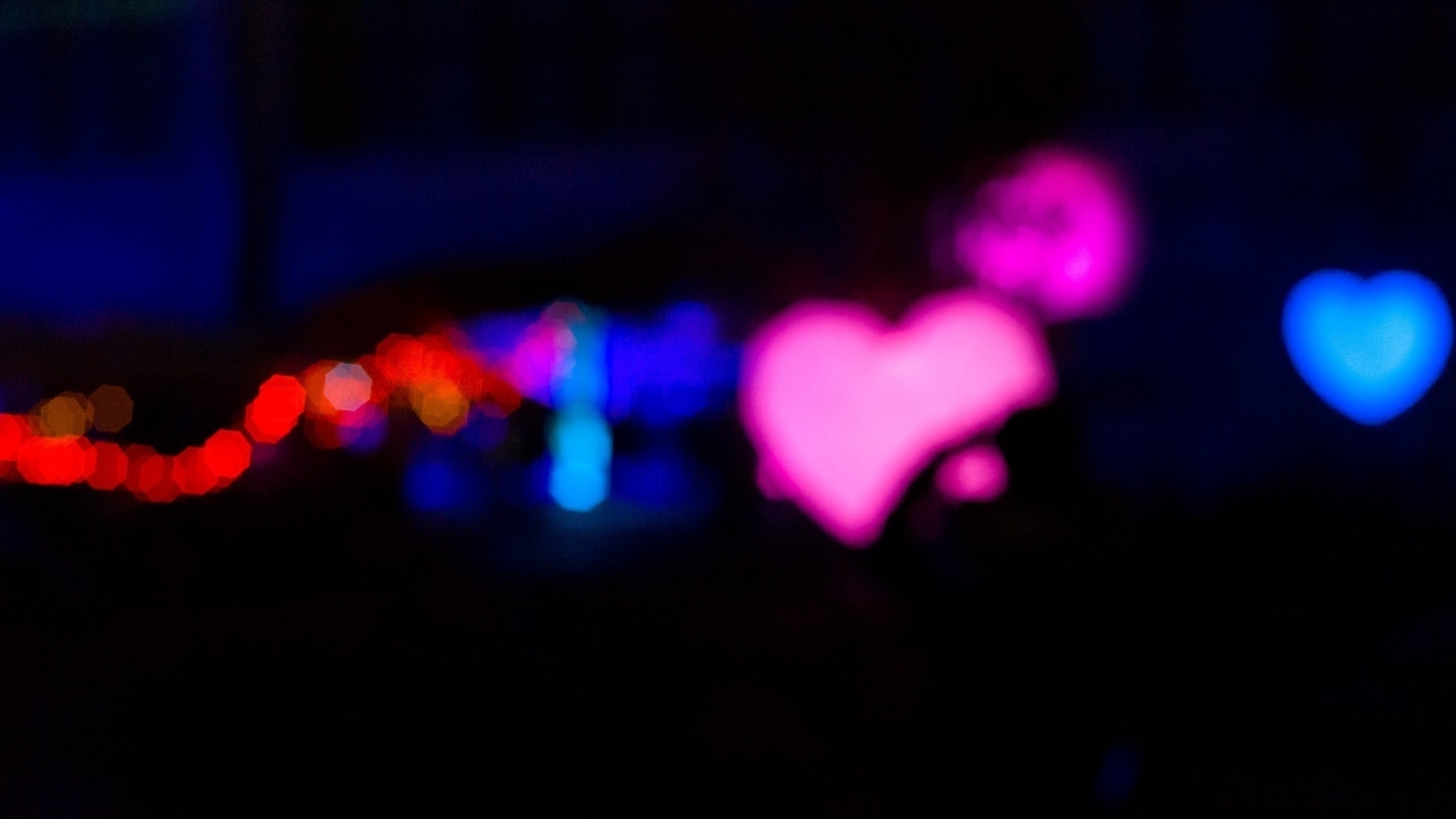 An out of focus image of blurry blobs of color and bokeh on a black background. A couple of the blobs resemble heart shapes.